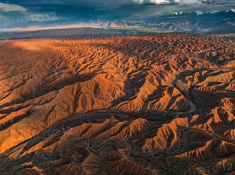 River bed runs through a landscape of eroded hills, badlands at sunset, mountain peaks of the Tian Shan Mountains in the background, aerial view, Canyon of the Forgotten Rivers, Issyk Kul, Kyrgyzstan, Asia