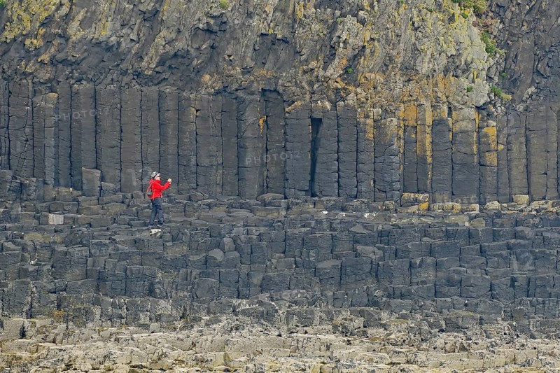 Person with red jacket standing in front of impressive basalt columns on a rocky cliff, Staffa, Mendelsons Hebrides Overture, Inner Hebrides, Scotland, United Kingdom, Europe