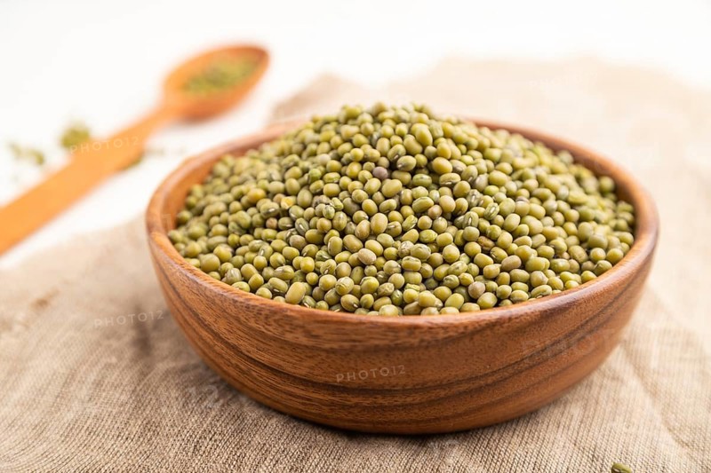 Wooden bowl with raw green mung bean and wooden spoon on a white background and linen textile. Side view, close up, selective focus