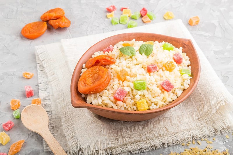 Bulgur porridge with dried apricots and candied fruits in clay bowl on a gray concrete background and linen textile. Side view, close up. Turkish traditional cuisine