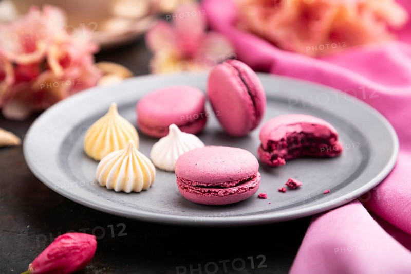 Purple macarons or macaroons cakes with cup of coffee on a black concrete background and pink textile. Side view, close up, selective focus
