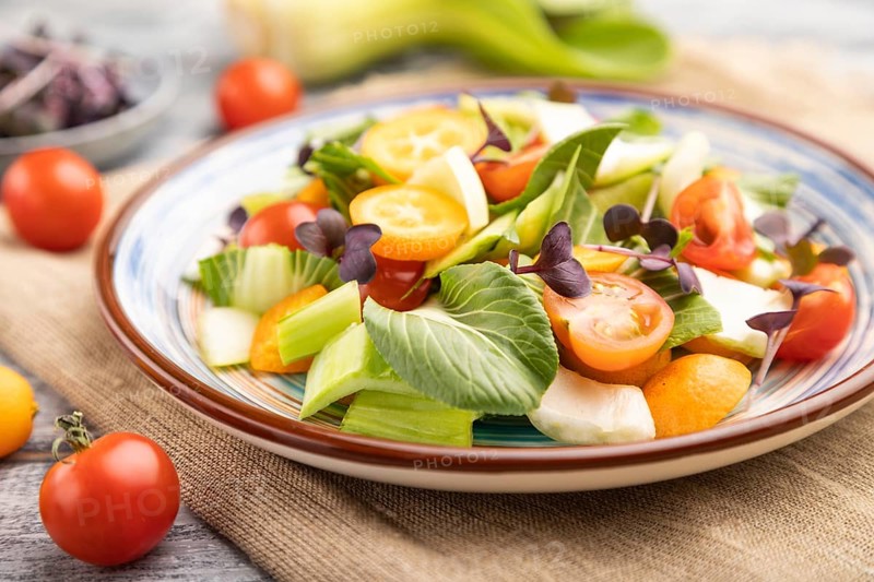 Vegetarian salad of pac choi cabbage, kiwi, tomatoes, kumquat, microgreen sprouts on gray wooden background and linen textile. Side view, close up, selective focus
