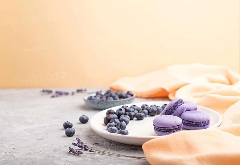 Purple macarons or macaroons cakes with blueberries on white ceramic plate on a gray and orange background and orange textile. Side view, copy space, selective focus