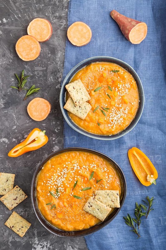 Sweet potato or batata cream soup with sesame seeds and snacks in blue ceramic bowls on a black concrete background with blue linen textile. top view, flat lay, close up