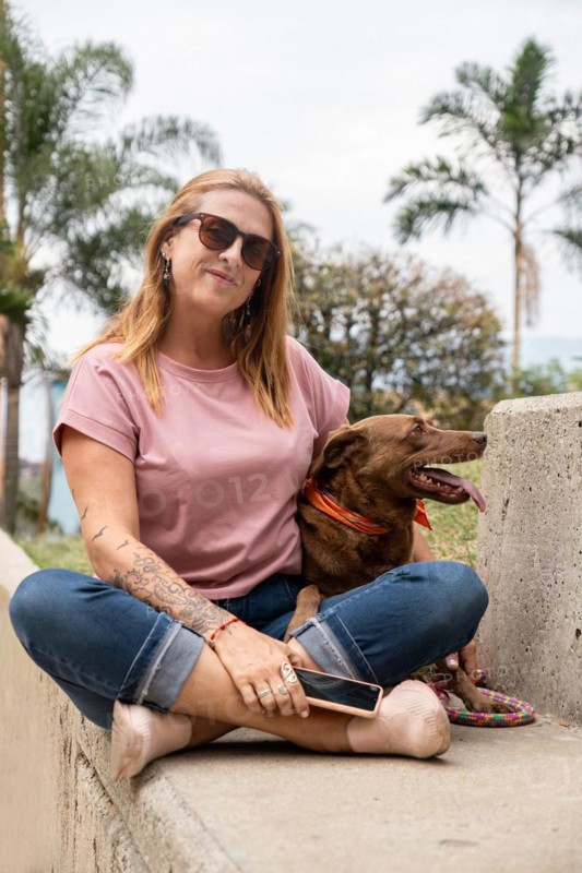 Middle-aged woman with sunglasses, sitting on a cement bench next to her dog, with palm trees in the background
