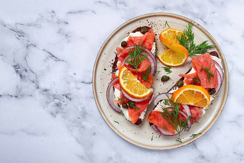 Sandwich with freshly smoked salmon, on black bread, with cottage cheese, with herbs and red onions, capers and pieces of citrus fruits, on a marble countertop, breakfast, no people