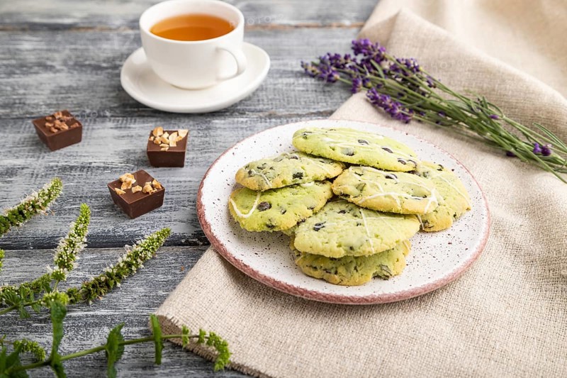 Green cookies with chocolate and mint on ceramic plate with cup of green tea and linen textile on gray wooden background. side view, close up