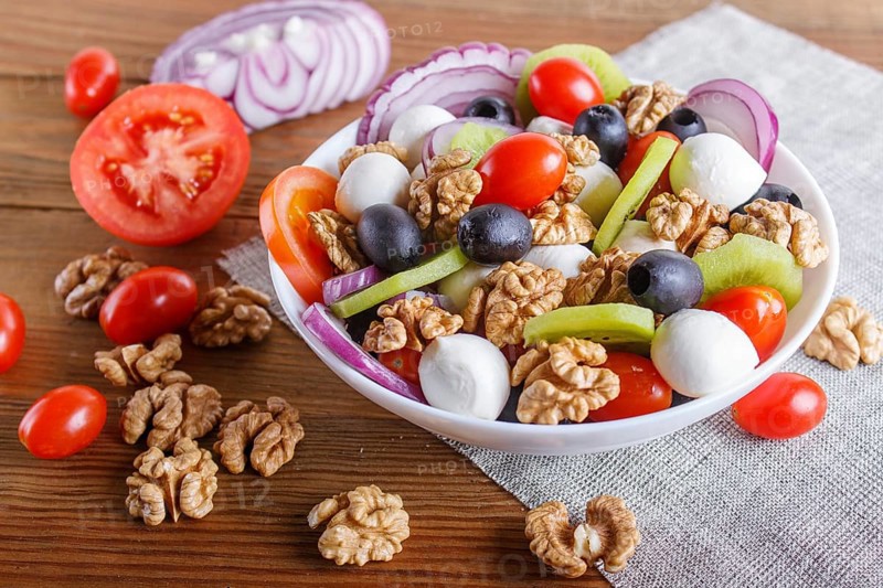 Salad with fresh cherry tomatoes, mozzarella cheese, olives, kiwi, onion and walnuts on brown wooden background, copy space, close up
