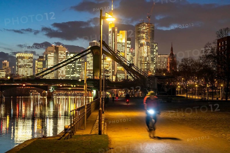 Skyline of the city centre of Frankfurt am Main, cyclist with light on the cycle path, pavement, promenade along the river Main, dusk, Hesse, Germany raft bridge in the background