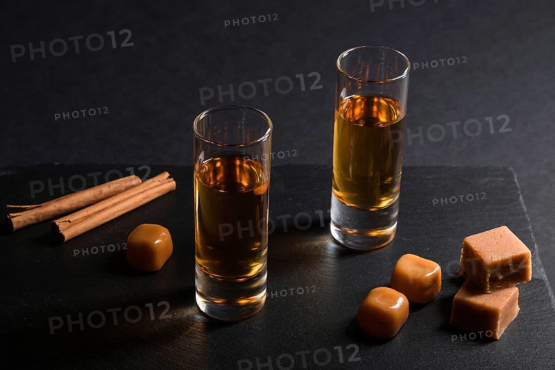 Glass of whiskey with caramel candies on a black stone slate board on black background. Side view, close up, low key concept