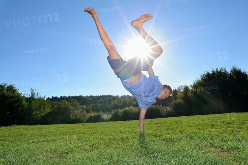 A boy doing a one-handed handstand on a grassy field with the sun shining brightly in the clear blue sky, Bavaria
