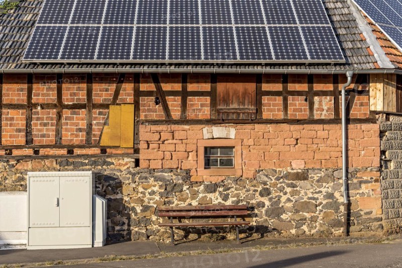 Historic farmhouse with photovoltaic system, solar panels on the roof, house wall with differently pieced masonry made of brick, sandstone, fieldstone and half-timbering, in front of it electricity distribution box and bench, Nidda, Hesse, Germany, Europe