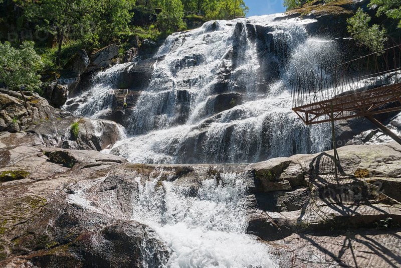 A large waterfall flows over rocks with a parapet in the foreground, surrounded by trees on a clear day, Cascada del Caozo, Garganta de Bonal de los Llanos, Piornal, Cáceres, Cacares, Extremadura, Spain, Europe