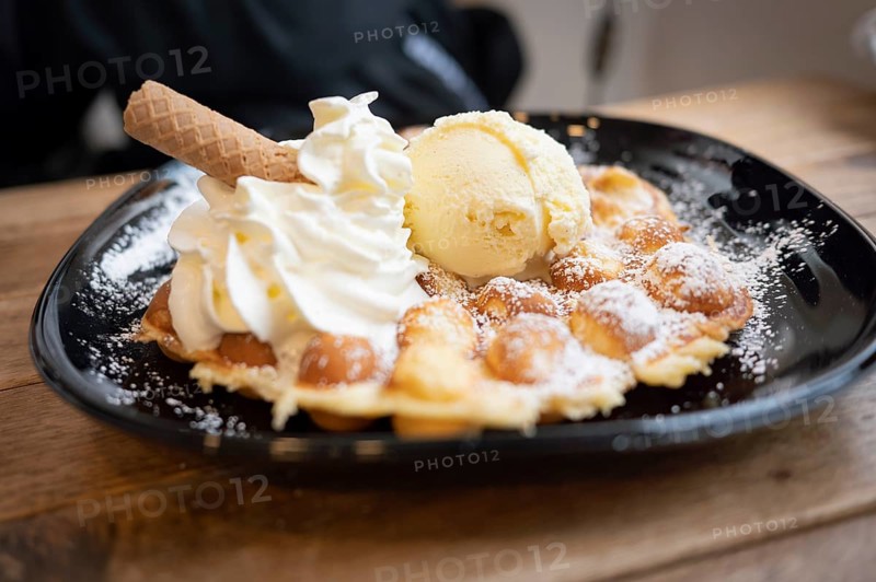 Belgian waffle with vanilla ice cream and whipped cream, waffle rolls, arranged on a black plate and dusted with icing sugar, on a wooden table, in the background a person dressed in black is recognisable, oblique frontal view, Wallonia, Belgium, Europe