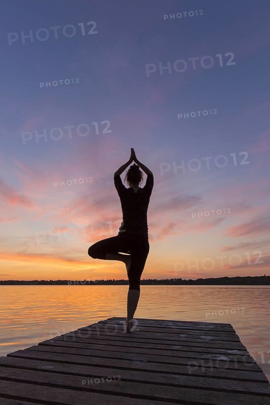Woman practicing standing posture Vrikshasana, Tree Pose, one legged balancing asana of the medieval hatha yoga on jetty at lake at sunset