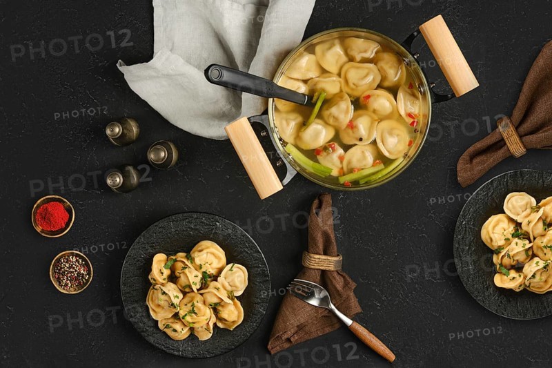 A pot filled with steaming dumplings sits on a dark textured surface. Two plates hold portions of dumplings, garnished with herbs and spices, inviting comfort and warmth for diners.