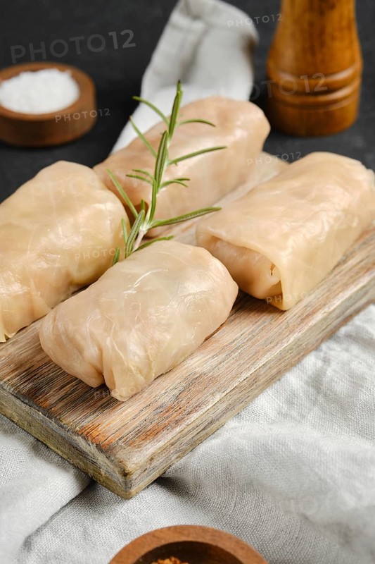 Close-up view of cabbage spring rolls stuffed with meat on a wooden board. A sprig of rosemary adds a touch of flavor, alongside salt on a surface, highlighting a healthy meal.