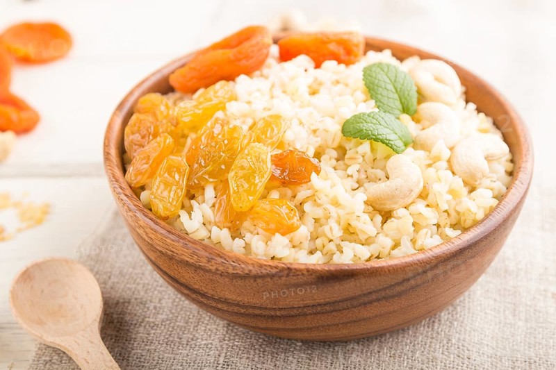 Bulgur porridge with dried apricots, raisins and cashew in wooden bowl on a white wooden background and linen textile. Side view, close up, selective focus. Turkish traditional cuisine