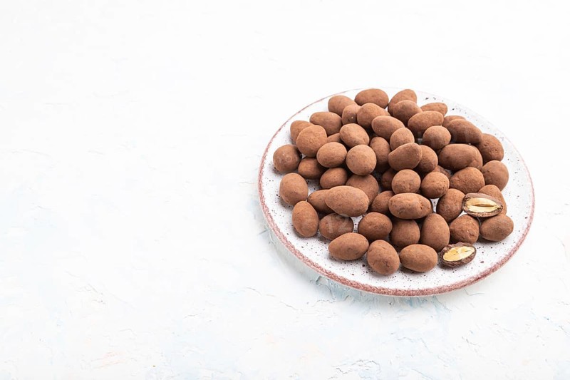 Almond in chocolate dragees on ceramic plate on white concrete background. Side view, copy space, close up