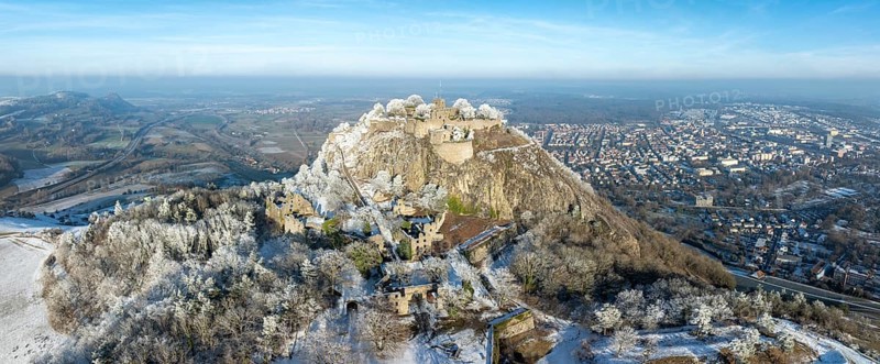 Aerial panorama of the snow-covered Hegau volcano Hohentwiel with Germany's largest castle ruins, surrounded by frosty trees in a winter landscape, with the town of Singen, district of Constance, Baden-Württemberg, Germany in the background
