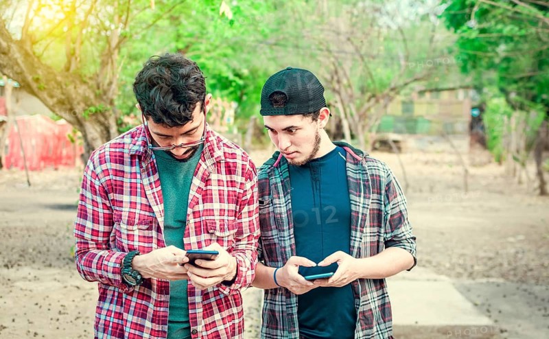 Jealous friend spying on his friend's cellphone outdoors, Friend spying on his friend phone. A guy snooping on his friend's phone on the street, Concept of jealous friends with cellphone