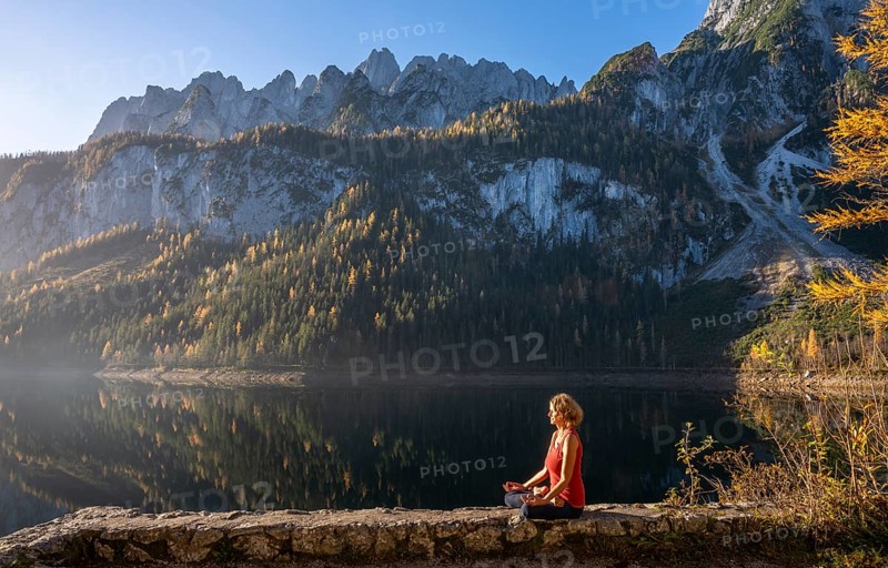 A woman doing yoga by the lake. Lotus position (Padmasana) . Vorderer Gosausee in autumn with a view of the Dachstein mountains. The Gosaukamm on the right. Blue sky, good weather. Reflection. Morning atmosphere. Vorderer Gosausee, Gosau, Gosau valley, Salzkammergut, Upper Austria, Austria, Europe