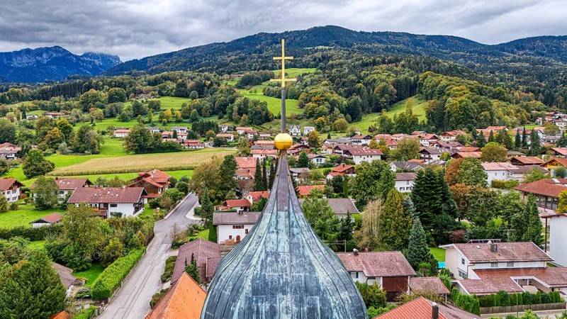 Village view with church spire cross, surrounded by green hills and wooded mountains, under a cloudy sky, church spire, Bad Feilnbach