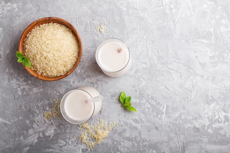 Organic non dairy rice milk in glass and wooden plate with rice seeds on a gray concrete background. Vegan healthy food concept, flat lay, top view, copy space