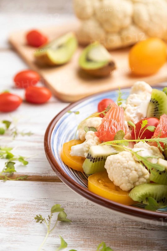 Vegetarian salad of cauliflower cabbage, kiwi, tomatoes, microgreen sprouts on white wooden background and blue linen textile. Side view, close up, selective focus