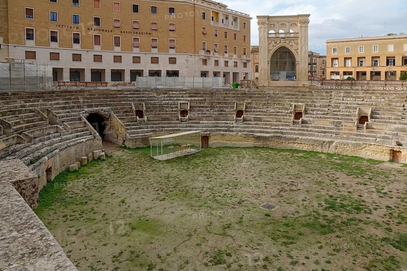 An ancient Roman amphitheatre made of stone with grass inside, surrounded by historic buildings, Lecce, Apulia, Italy, Europe