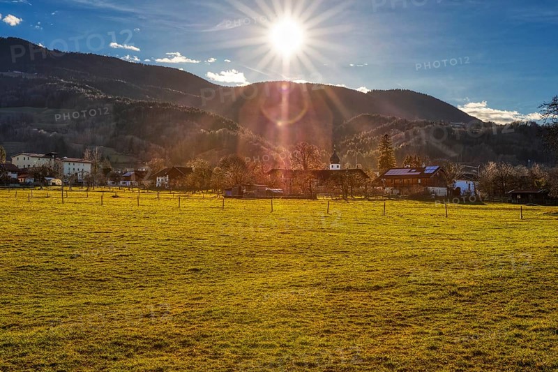 The sun sets over a village that lies in front of a mountain range and is surrounded by green fields, Bad Feilnbach