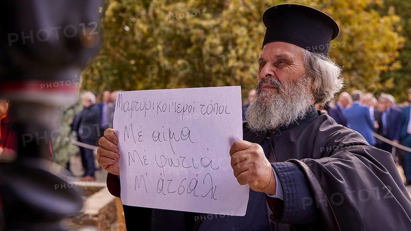 Priest holding a sign in the midst of a crowd in autumnal surroundings, Visit of the Federal President Frank-Walter Steinmeier in Kandanos on 31 October 2024, Federal President, Frank-Walter Steinmeier, memorial, war crimes, Nazis, Wehrmacht crimes, World War II, Kandanos, Southwest Crete, Crete, Greek Islands, Greece, Europe