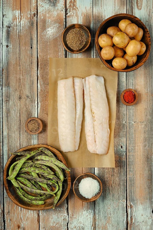 Two fresh cod fish fillets on parchment paper surrounded by various spices, green beans, and small potatoes on a wooden table, showcasing a vibrant kitchen arrangement perfect for cooking