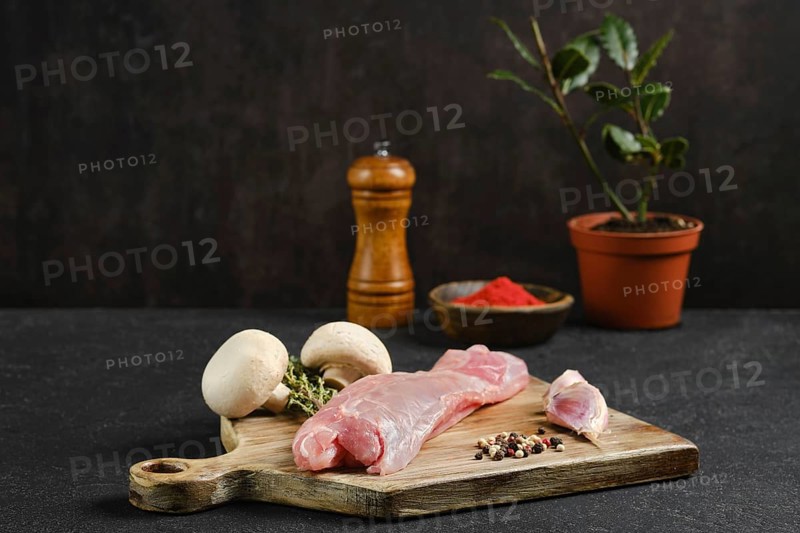 Raw rabbit tenderloin on a wooden cutting board beside fresh mushrooms and an assortment of spices. A pepper mill and a potted herb add a rustic touch to this culinary setup, inviting cooking.