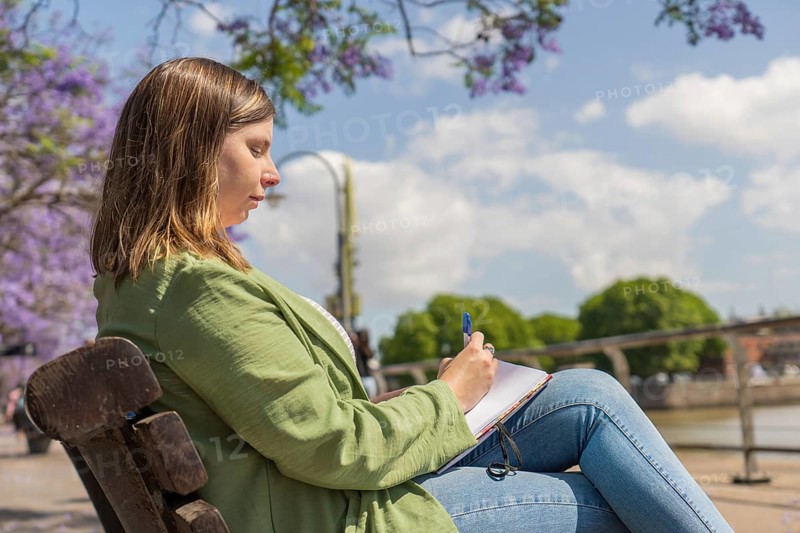 A young woman sitting on a bench in a park, writing in a notebook with a pen. The background features trees, flowers, and a sunny sky, creating a peaceful and creative atmosphere