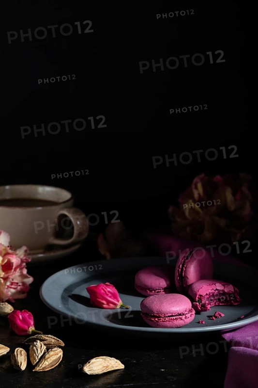 Purple macarons or macaroons cakes with cup of coffee on a black concrete background and pink textile. Hard light, low key. Side view, copy space