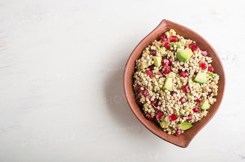 Salad of germinated buckwheat, avocado, walnut and pomegranate seeds in clay plate on white wooden background. Top view, copy space