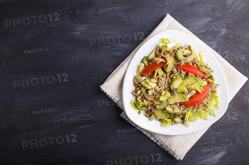 Vegetarian salad of celery, germinated rye, tomatoes and avocado on linen tablecloth, top view, copy space, black background