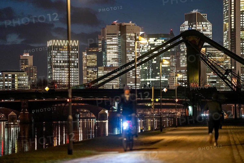 Skyline of the city centre of Frankfurt am Main, cyclist with light on the cycle path, pavement, promenade along the river Main, dusk, Hesse, Germany raft bridge in the background