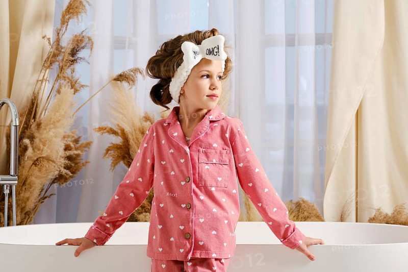 Cute child in a pink pajama and hair band stands next to a modern white bathtub spreading hands aside and looks to the side