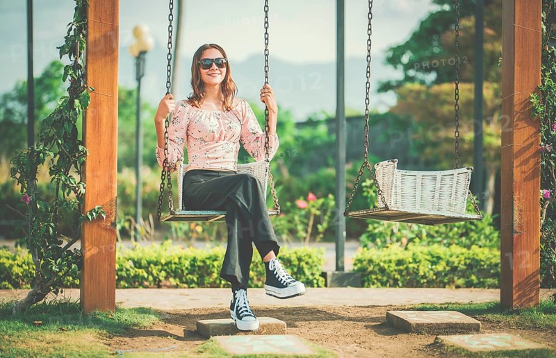 Cheerful girl in glasses relaxing on a swing in a natural park. Happy young woman sitting on a swing in a nature park