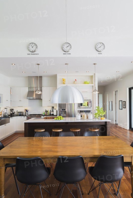Open-concept dining and kitchen area with antique dining table with black leather and plastic chairs, marble top wooden island with painted black steel and exotic wood bar stools, white cabinets inside a luxurious modern cube style house, Quebec, Canada, North America