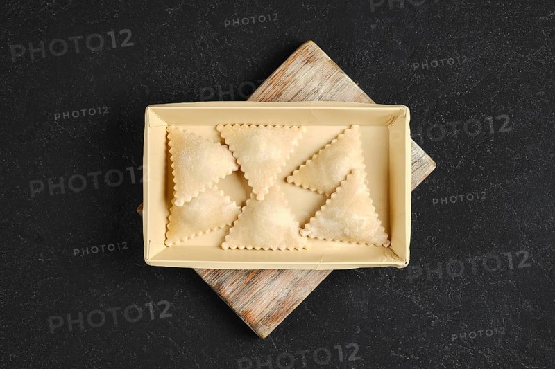 Freshly made ravioli are arranged neatly in a rectangular tray on a rustic wooden board. Each pasta piece is shaped with precise edges, showcasing a smooth texture.