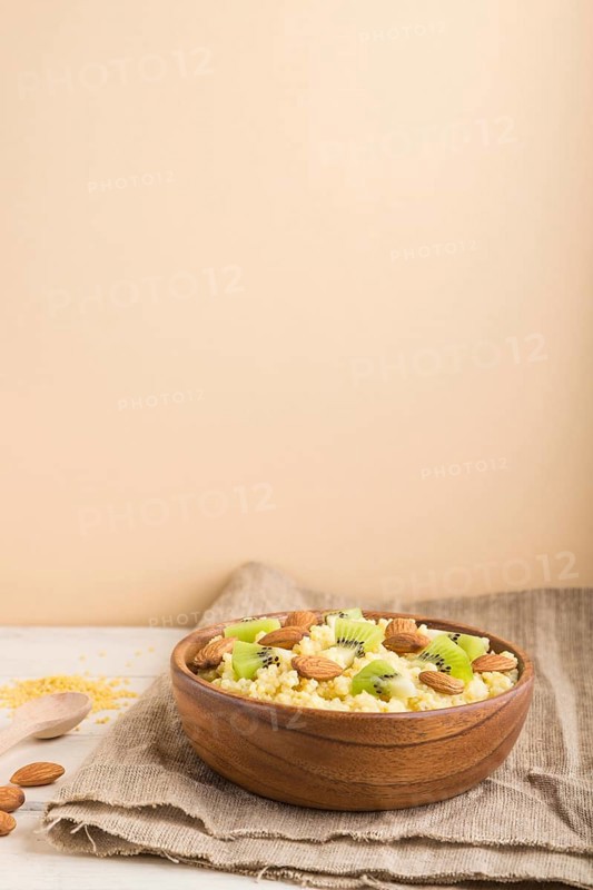 Millet porridge with kiwi and almonds in wooden bowl on a white and orange background and linen textile. Side view, copy space, selective focus