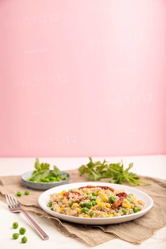 Quinoa porridge with green pea, corn and dried tomatoes on ceramic plate on a white and pastel pink background and linen textile. Side view, copy space
