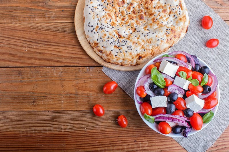 Greek salad with fresh cherry tomatoes, feta cheese, black olives, basil and onion on wooden background, pancake, top view, copy space