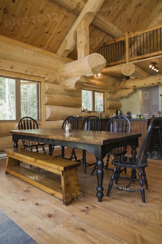 Black stained antique wood dining table with rounded high back chairs and pine wood sitting bench in open concept dining room and kitchen inside handcrafted Eastern white pine Scandinavian log cabin home, Quebec, Canada, North America