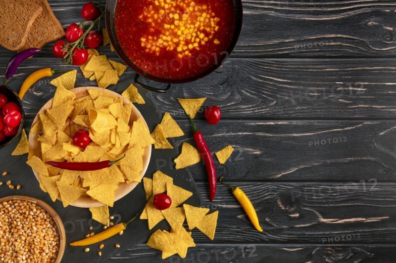 Tortilla chips next to a pan with chilli con carne, peppers and tomatoes on a dark wooden background