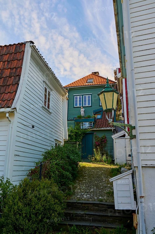 Narrow alley with cobblestones between historic houses, foreground white buildings, background green house with red tiled roof and lantern in daylight, Bergen, Vestland, Norway, Europe