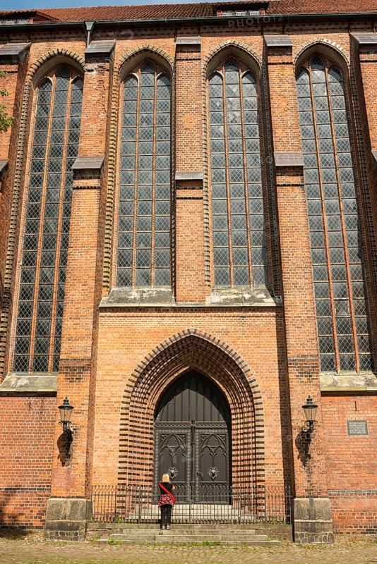 Large brick church with high windows, pointed arches, a person stands at the entrance, historic wooden door with lanterns, facade of St Michael's Church, sunny day in summer, Old Town of the Hanseatic City of Lüneburg, Lower Saxony, Germany, Europe